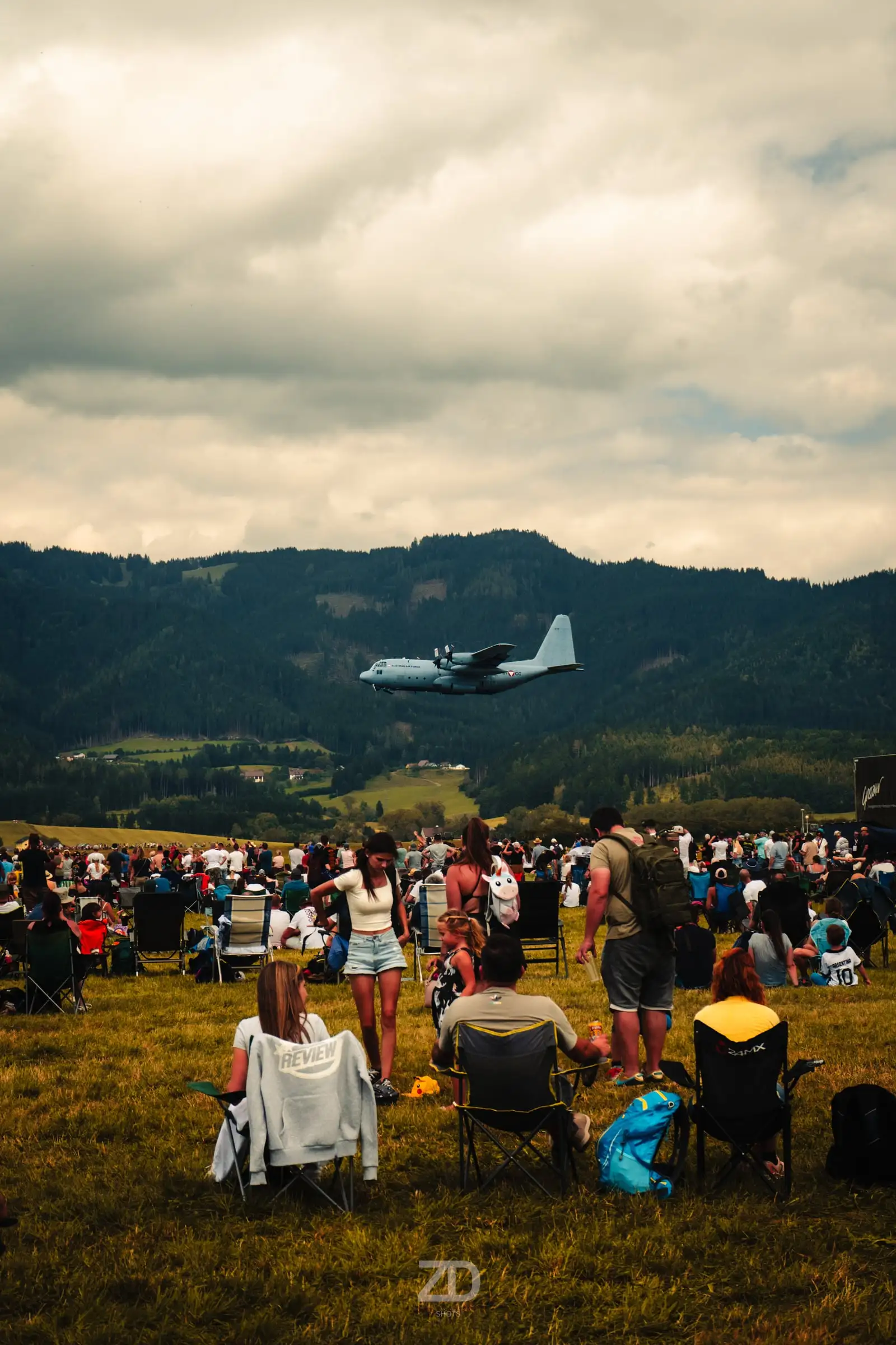 Publikum auf einer Wiese, darüber ein Flugzeug vor Bergen