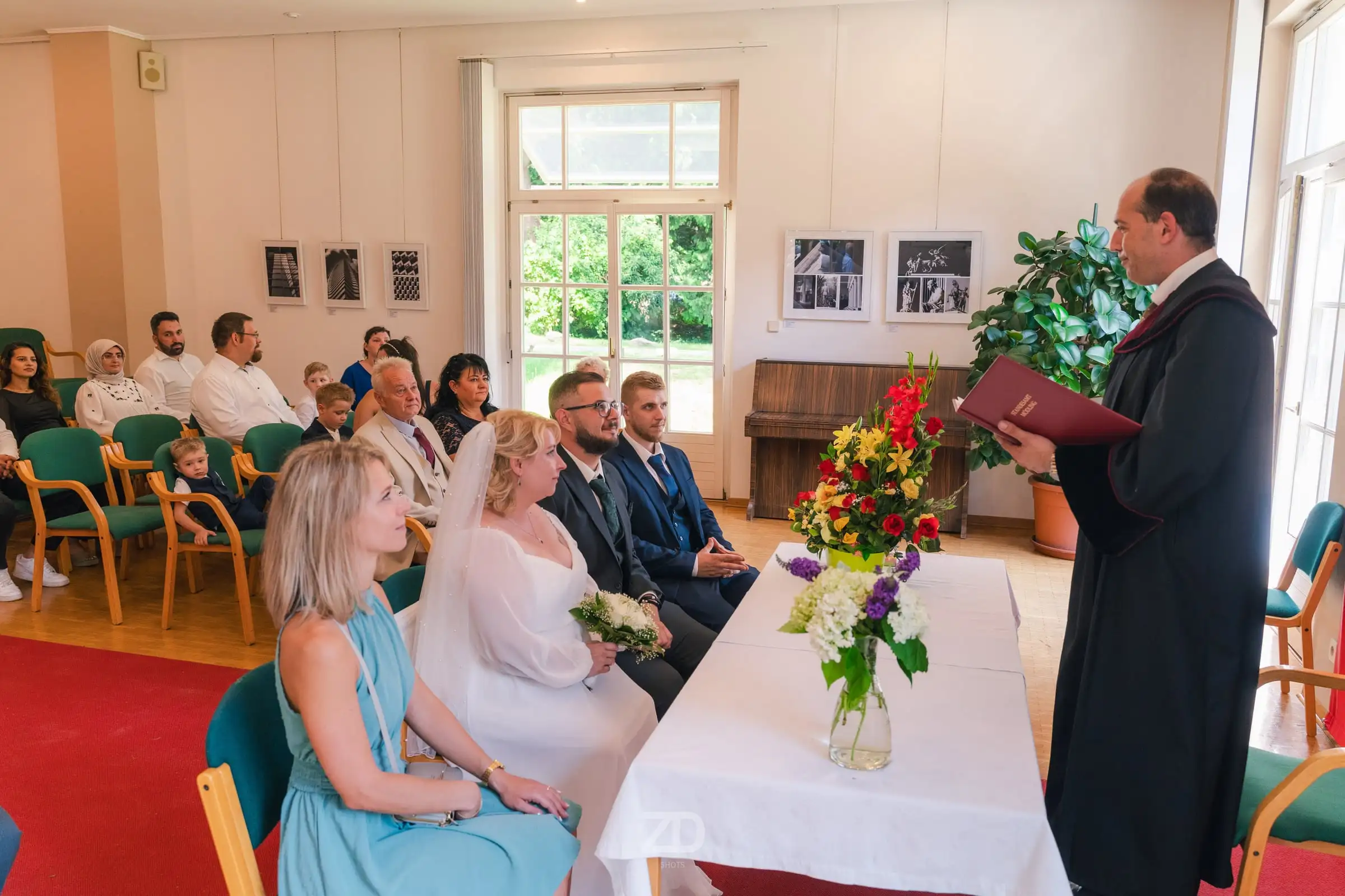 Trauung im Saal: Das Brautpaar sitzt vor dem Blumentisch, die Gäste dahinter