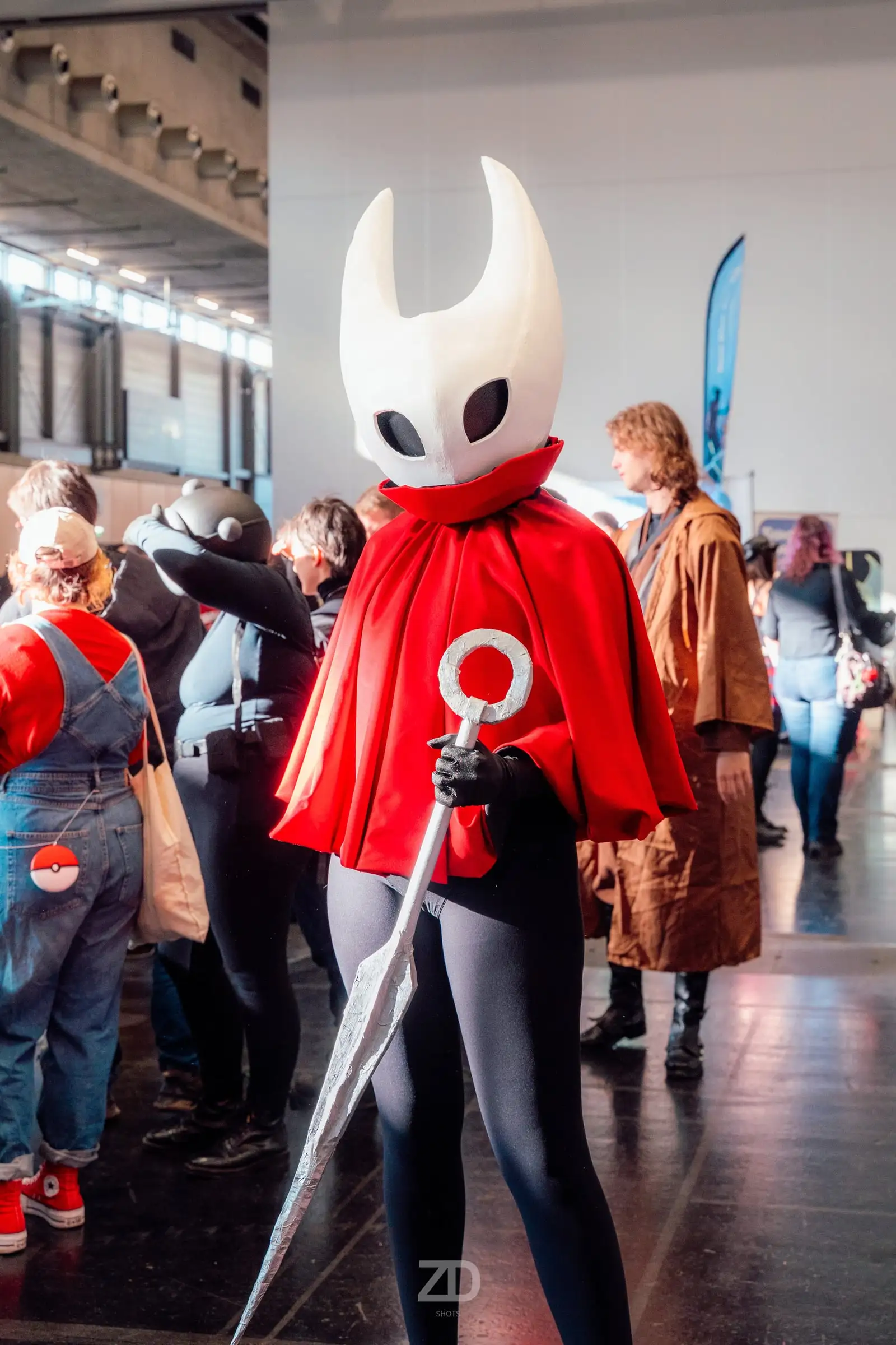 Cosplayerin mit weißer Maske und rotem Umhang in einer Messehalle